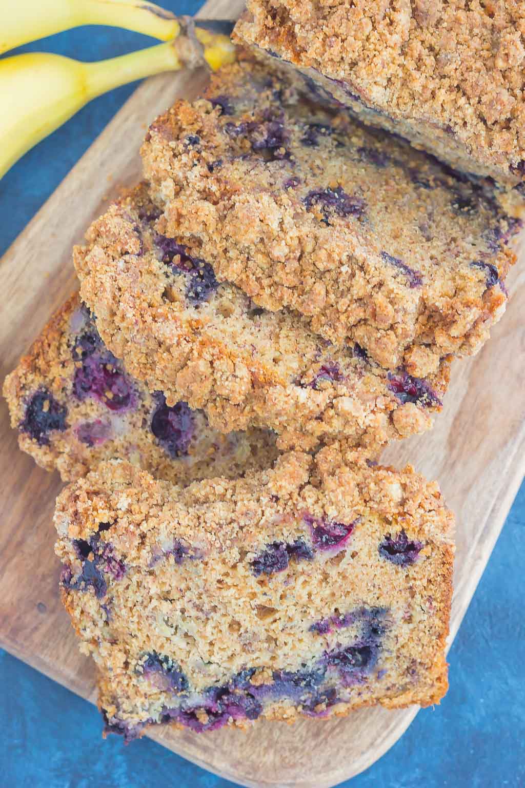 overhead view of blueberry banana bread on wood cutting board next to bunch of bananas