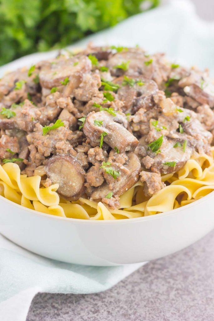 A bowl of easy ground beef stroganoff features egg noodles topped with a creamy mixture of ground beef, sliced mushrooms, and fresh parsley. The dish rests on a light surface with a softly blurred green background.