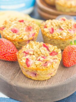 Three baked oatmeal cups with visible oats and strawberry pieces are displayed on a round wooden board, alongside fresh strawberries for a delightful touch.
