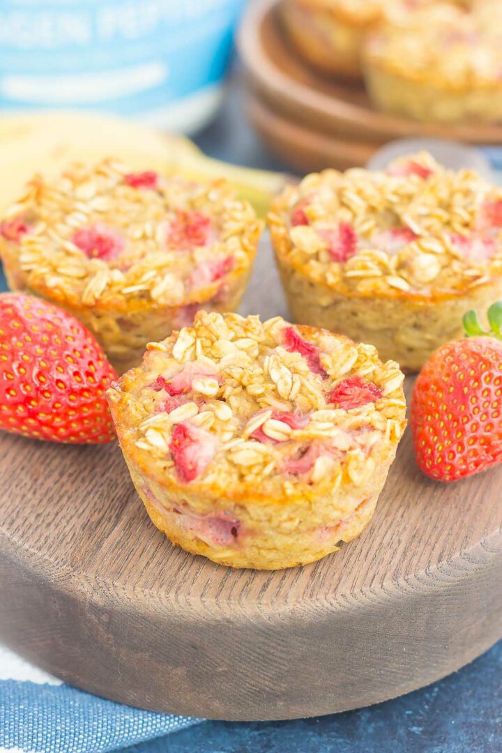 Three baked oatmeal cups with visible oats and strawberry pieces are displayed on a round wooden board, alongside fresh strawberries for a delightful touch.