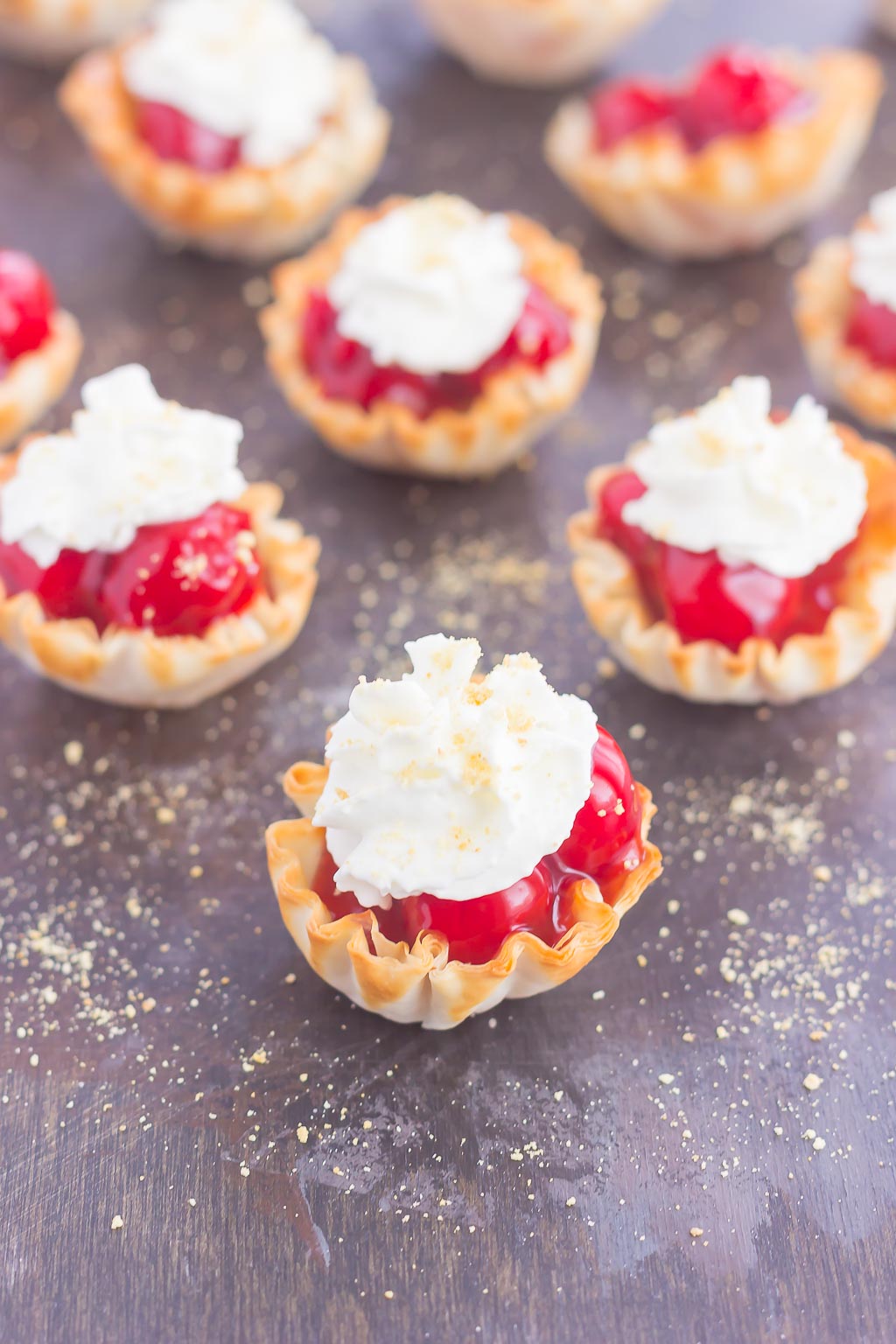 mini cherry pie bites topped with whipped cream on a gray surface. 