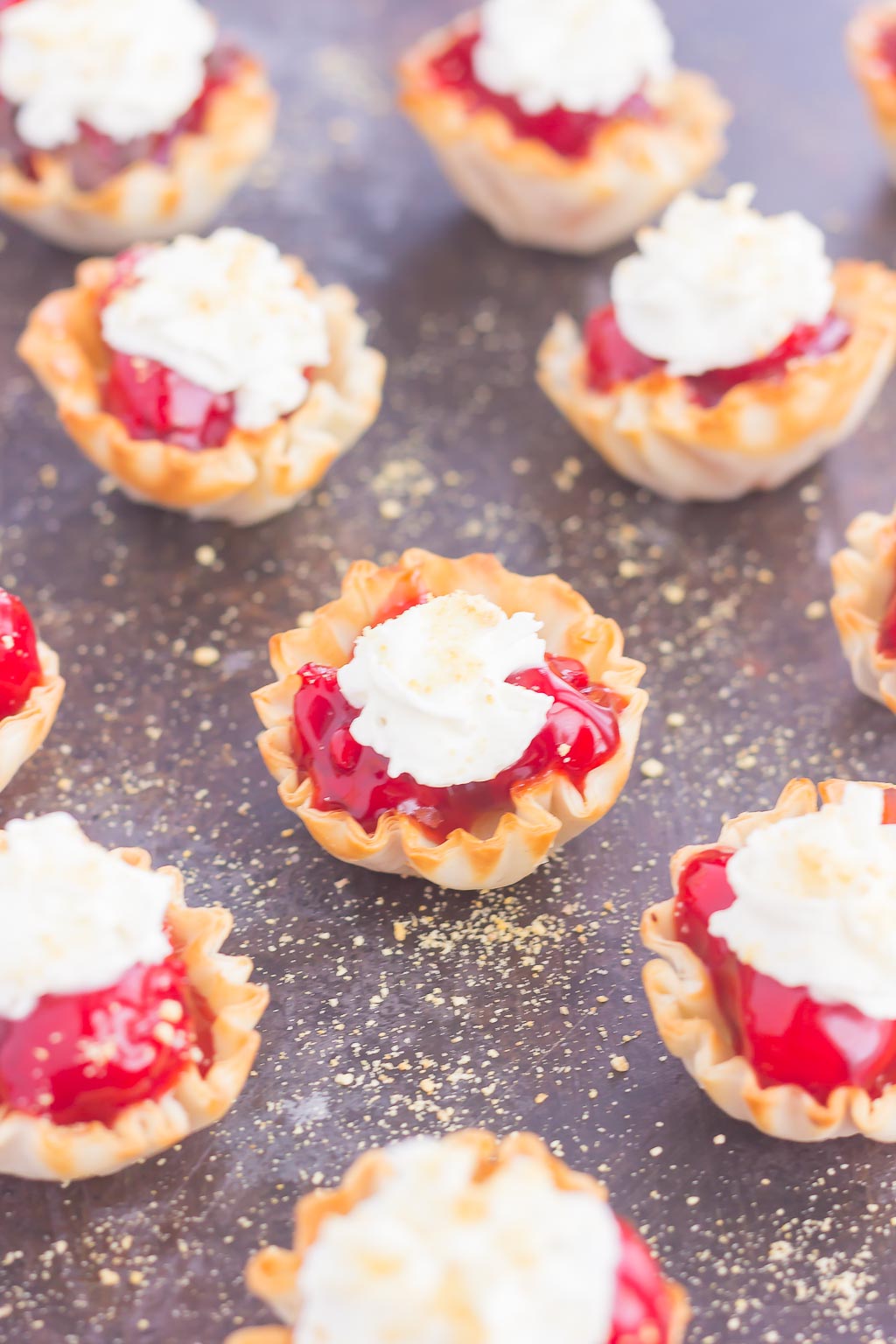 mini cherry pie bites topped with whipped cream on a gray surface. 