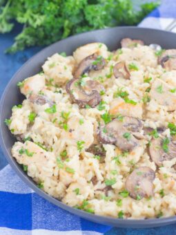 A skillet filled with creamy one pan chicken and mushroom parmesan rice, featuring sliced mushrooms, juicy chicken, and fresh parsley, sits on a blue and white checkered cloth with parsley in the background.