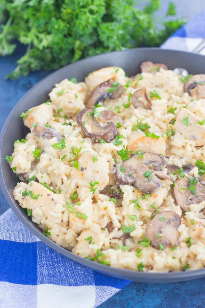 A skillet filled with creamy one pan chicken and mushroom parmesan rice, featuring sliced mushrooms, juicy chicken, and fresh parsley, sits on a blue and white checkered cloth with parsley in the background.
