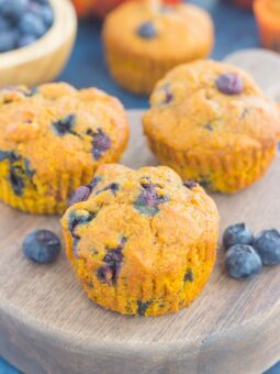 Three pumpkin blueberry muffins are displayed on a wooden board, with fresh blueberries scattered around and more muffins and fruit visible in the blurred background.