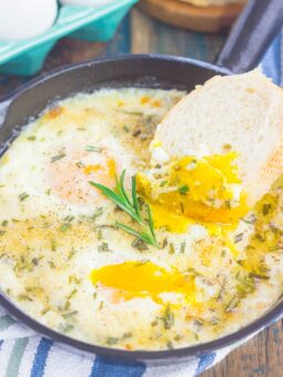 A skillet with oven baked eggs topped with herbs, one yolk broken and runny. A piece of bread is dipped into the yolk, with a sprig of rosemary on top. Eggs and bread are visible in the background.