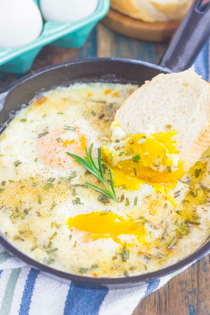 A skillet with oven baked eggs topped with herbs, one yolk broken and runny. A piece of bread is dipped into the yolk, with a sprig of rosemary on top. Eggs and bread are visible in the background.