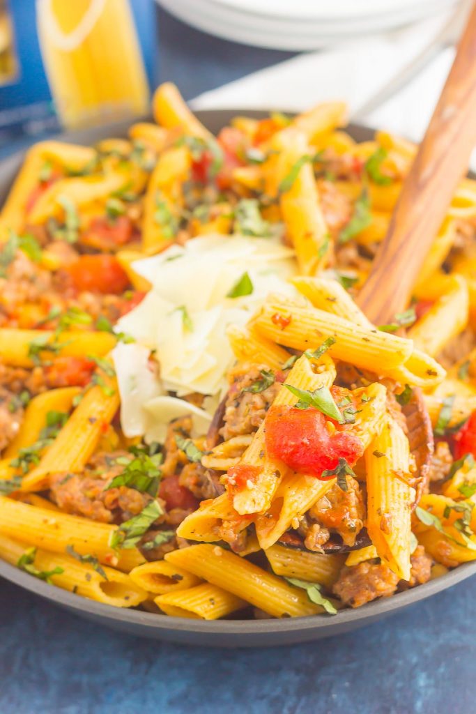 A close-up of a pan filled with penne, italian sausage pasta, tomatoes, fresh herbs, and topped with shaved parmesan cheese. A wooden spoon lifts a serving of the pasta above the pan.