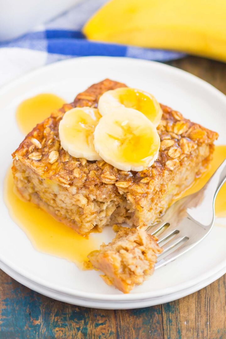 A square slice of banana baked oatmeal topped with banana slices and syrup sits on a white plate. A fork with a bite-sized piece rests beside it, while a banana is blurred in the background.