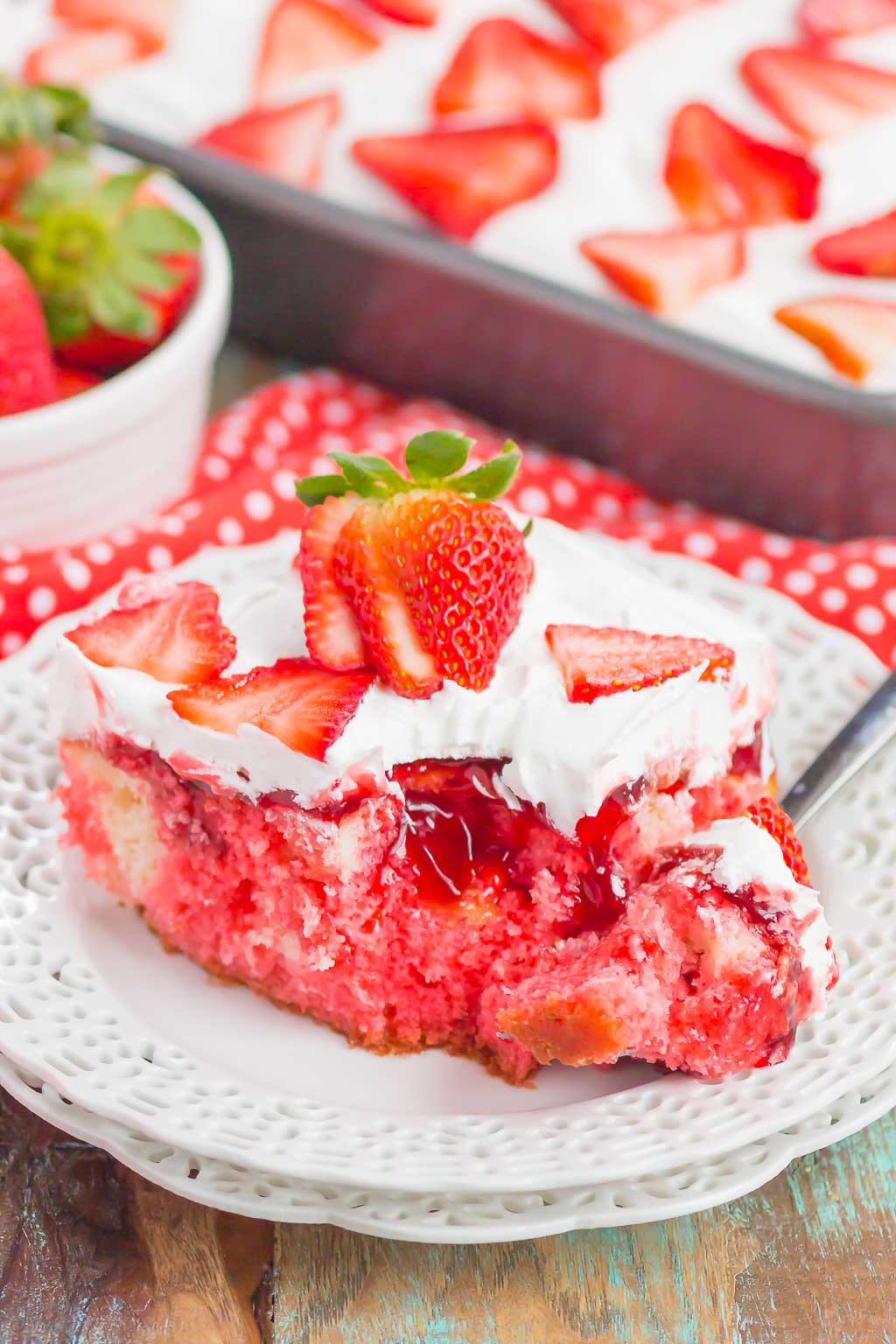 A strawberry poke cake slice on a white plate. A bite has been speared on a fork, which is resting on the plate. 
