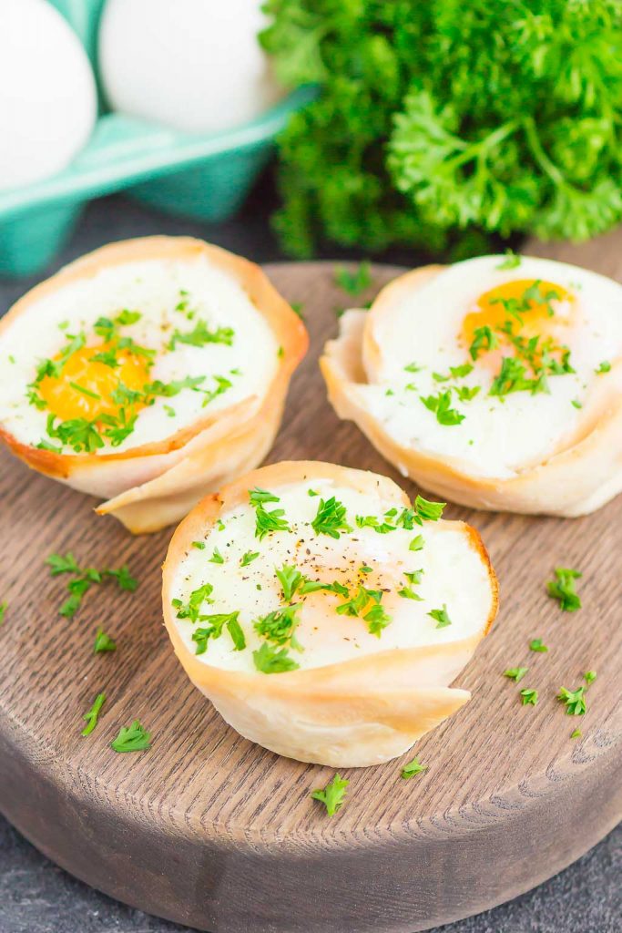 Three turkey egg cups garnished with chopped parsley sit on a round wooden board, with fresh parsley and eggs in the background.