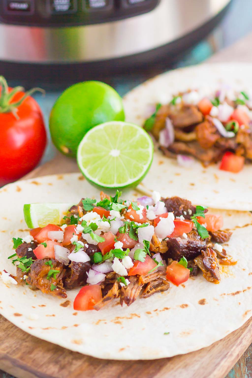 Two bbq pork tacos lying flat to show fillings. Lime halves and fresh tomato rest in the background. 