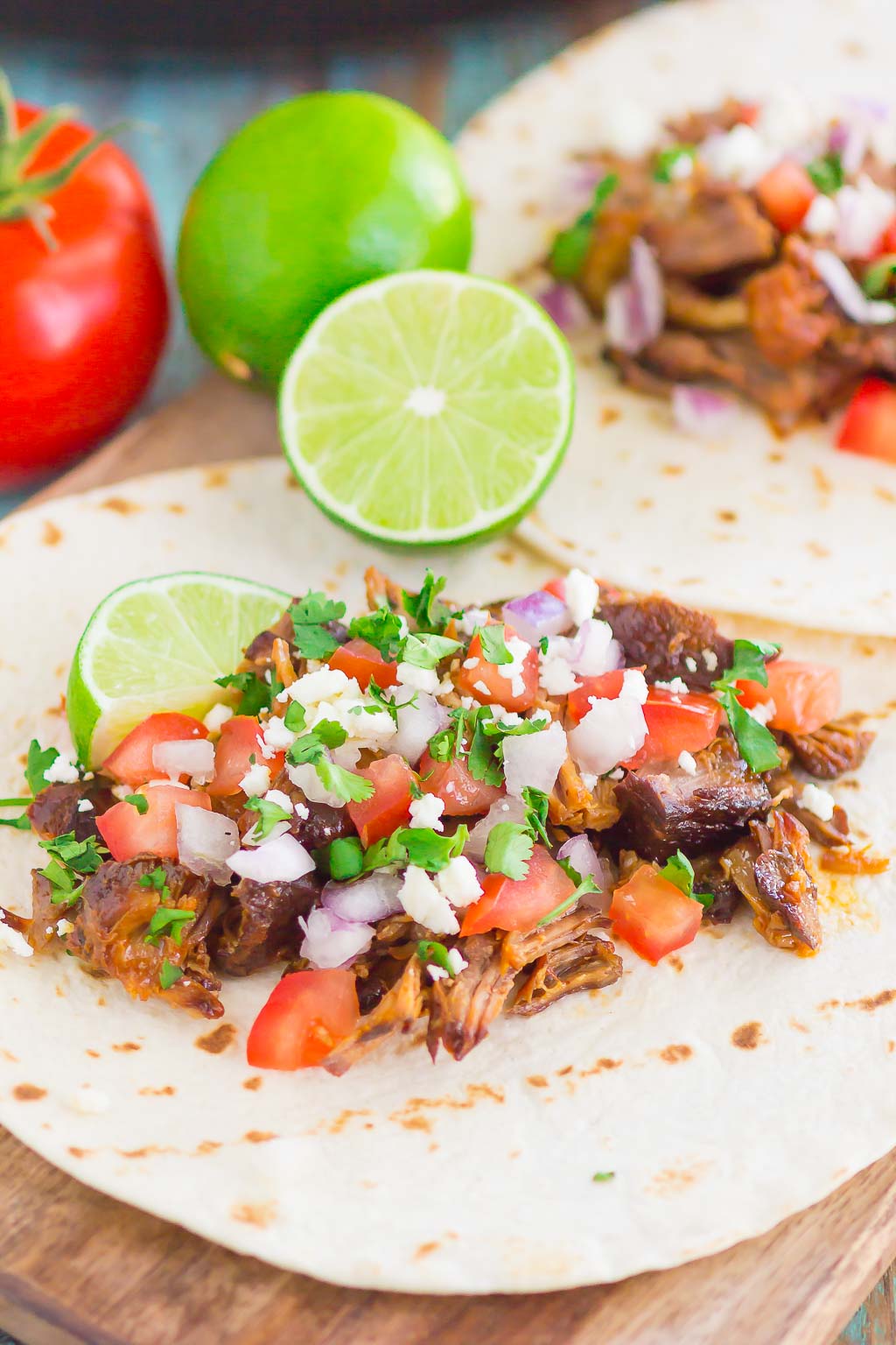 Two bbq pork tacos lying flat to show fillings. Lime halves and fresh tomato rest in the background. 