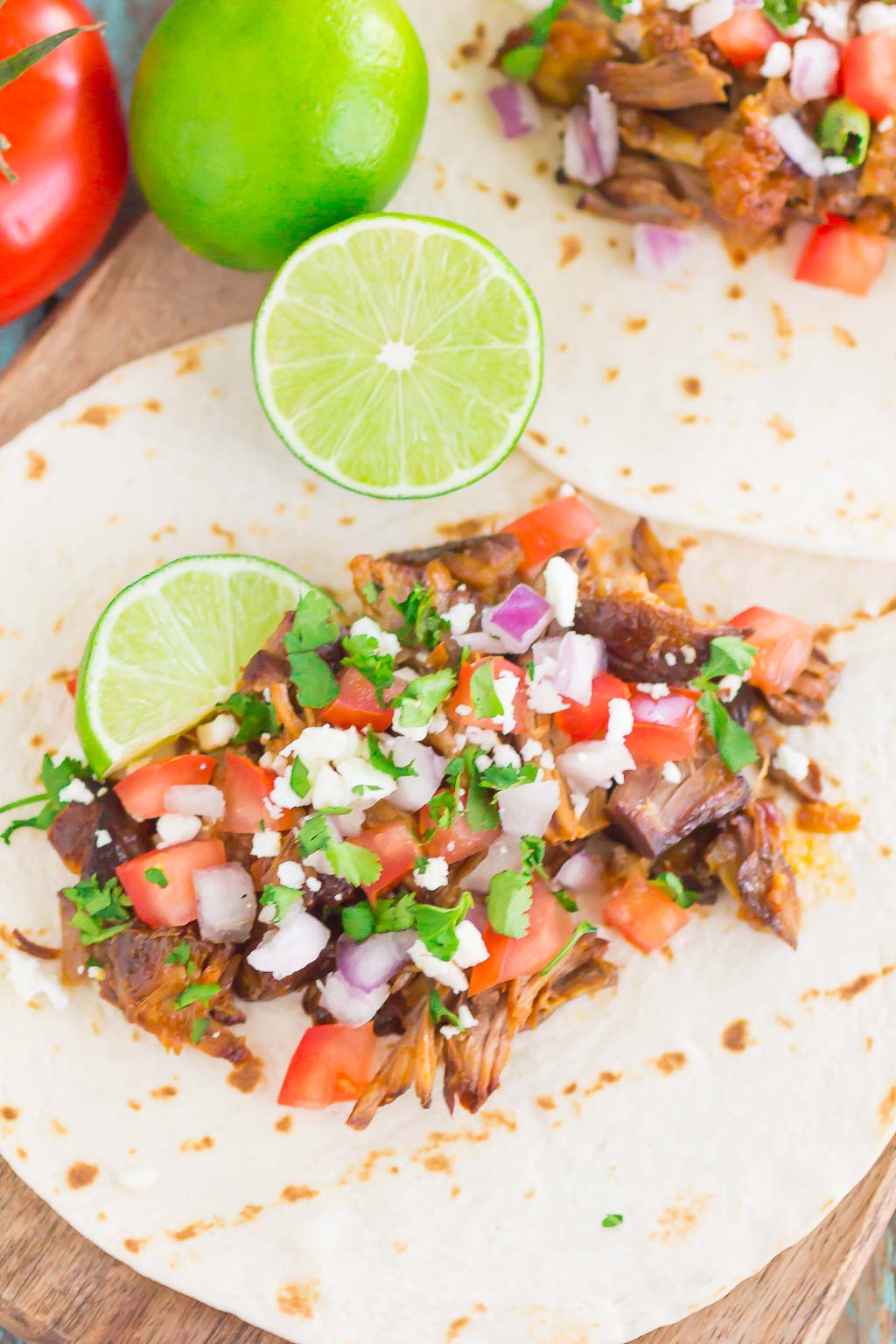 Two bbq pork tacos lying flat to show fillings. Lime halves and fresh tomato rest in the background. 