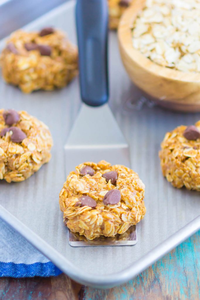 A metal spatula lifts an almond butter oatmeal cookie with chocolate chips from a baking sheet. Other cookies and a wooden bowl of oats are visible in the background. The cookies look soft and chewy.