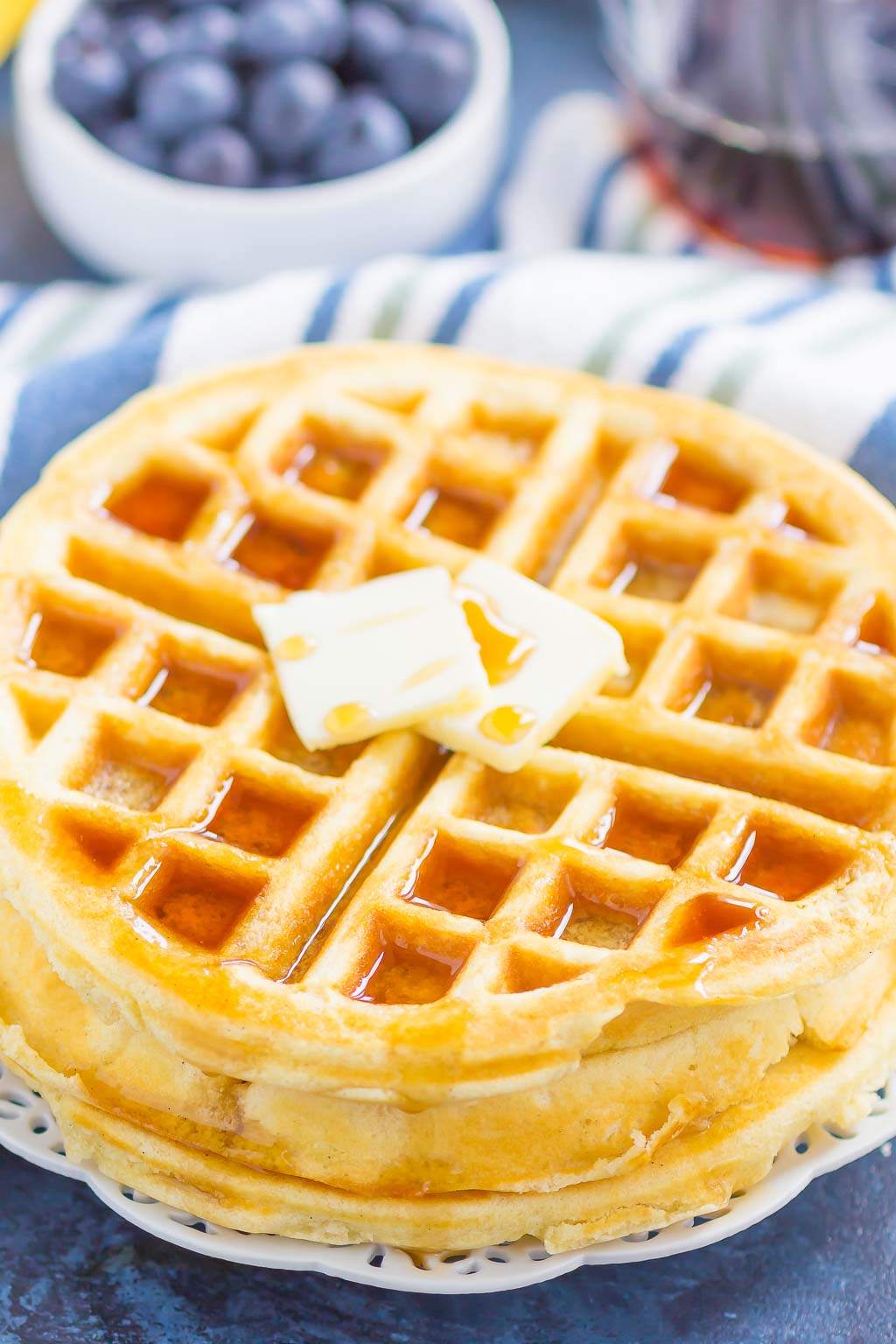 A stack of fluffy Belgian waffles topped with butter and maple syrup. A bowl of blueberries rests in the background. 