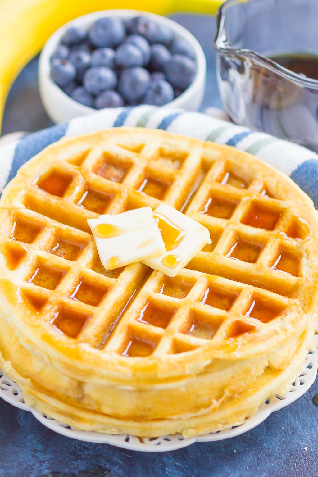 A stack of fluffy Belgian waffles topped with butter and maple syrup. A bowl of blueberries rests in the background. 