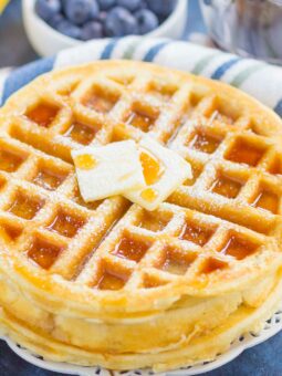 A close-up of fluffy Belgian waffles stacked high, topped with two pats of butter and drizzled with syrup, with a bowl of blueberries and a striped napkin in the background.