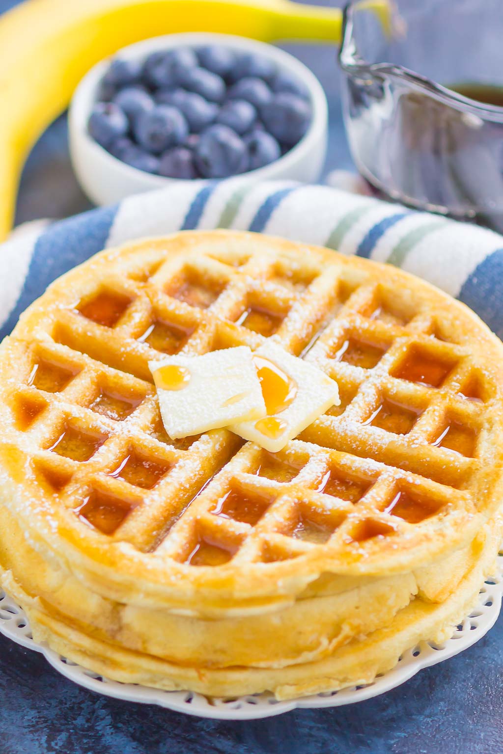 A stack of fluffy Belgian waffles topped with butter and maple syrup. A bowl of blueberries rests in the background. 