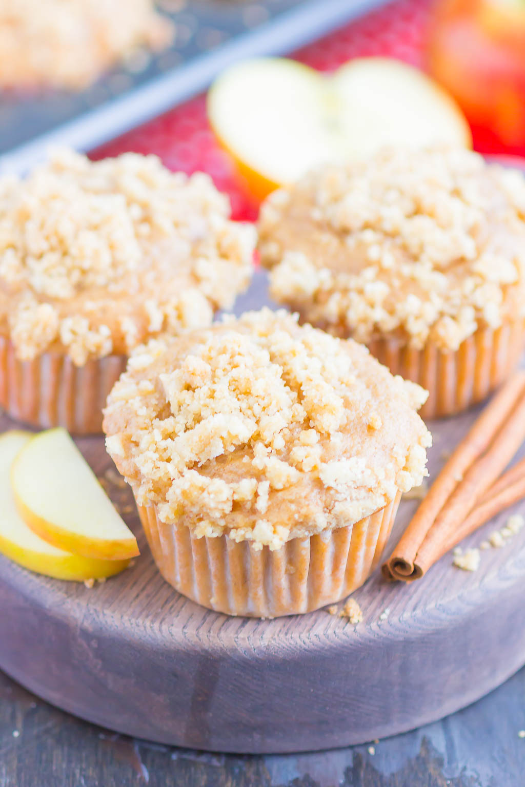 three apple streusel muffins  on a wood tray with cinnamon sticks and apple slices 