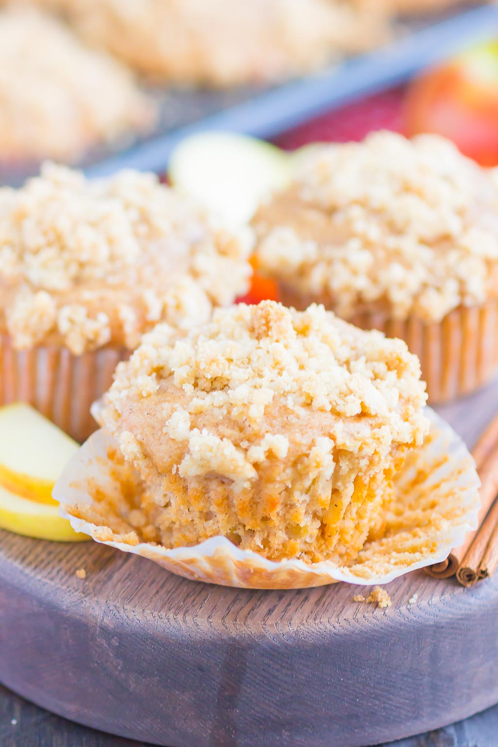 three apple cinnamon muffins on a wood tray. the front muffin has the paper liner pulled down. 