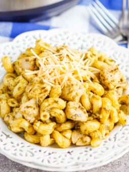A white decorative plate filled with instant pot pesto chicken pasta, featuring cavatappi and chunks of chicken in creamy pesto sauce, topped with shredded parmesan cheese. Forks are visible in the background.