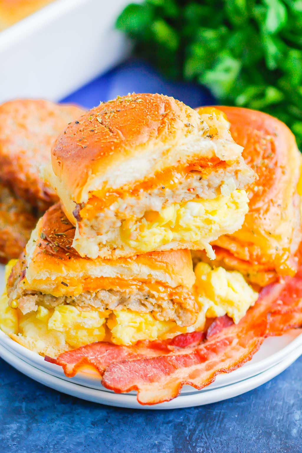 A plate with two breakfast sliders filled with scrambled eggs, sausage, and cheese, stacked on each other, served alongside crispy bacon strips. Breakfast sliders steal the show as fresh greens blur in the background.