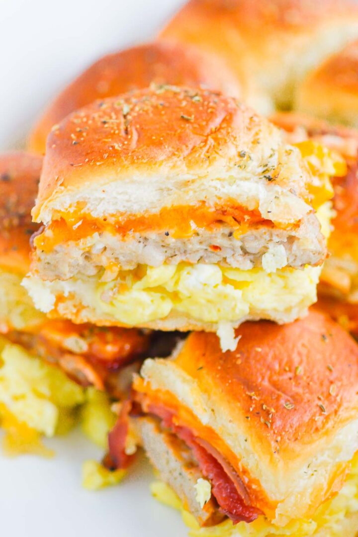 A close-up of breakfast sliders made with golden buns, scrambled eggs, sausage, cheese, and bacon, stacked and cut to showcase the mouthwatering layers inside these breakfast sliders.