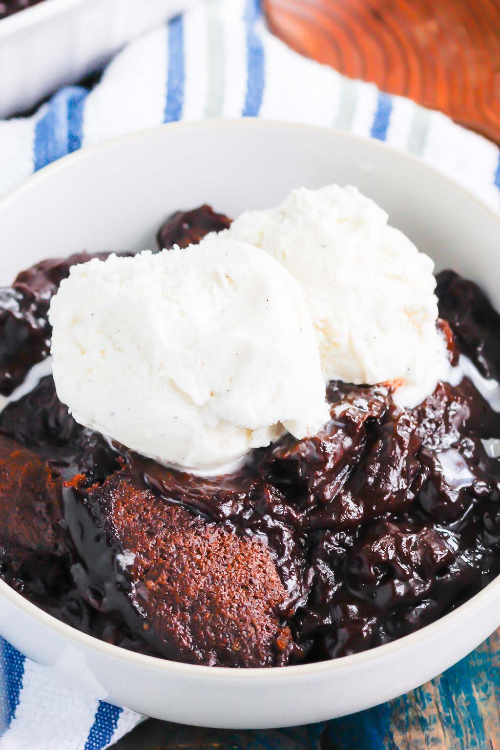 serving of chocolate cobbler topped with ice cream in a white bowl. a wooden spoon rests in the background. 