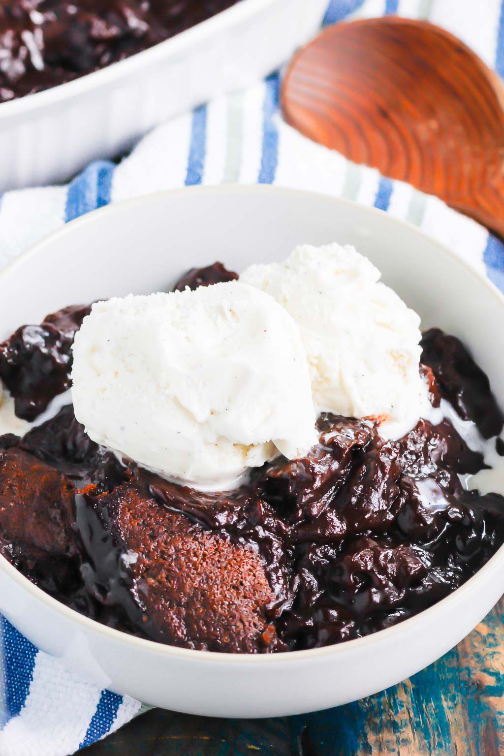 serving of chocolate cobbler topped with ice cream in a white bowl. a wooden spoon rests in the background. 