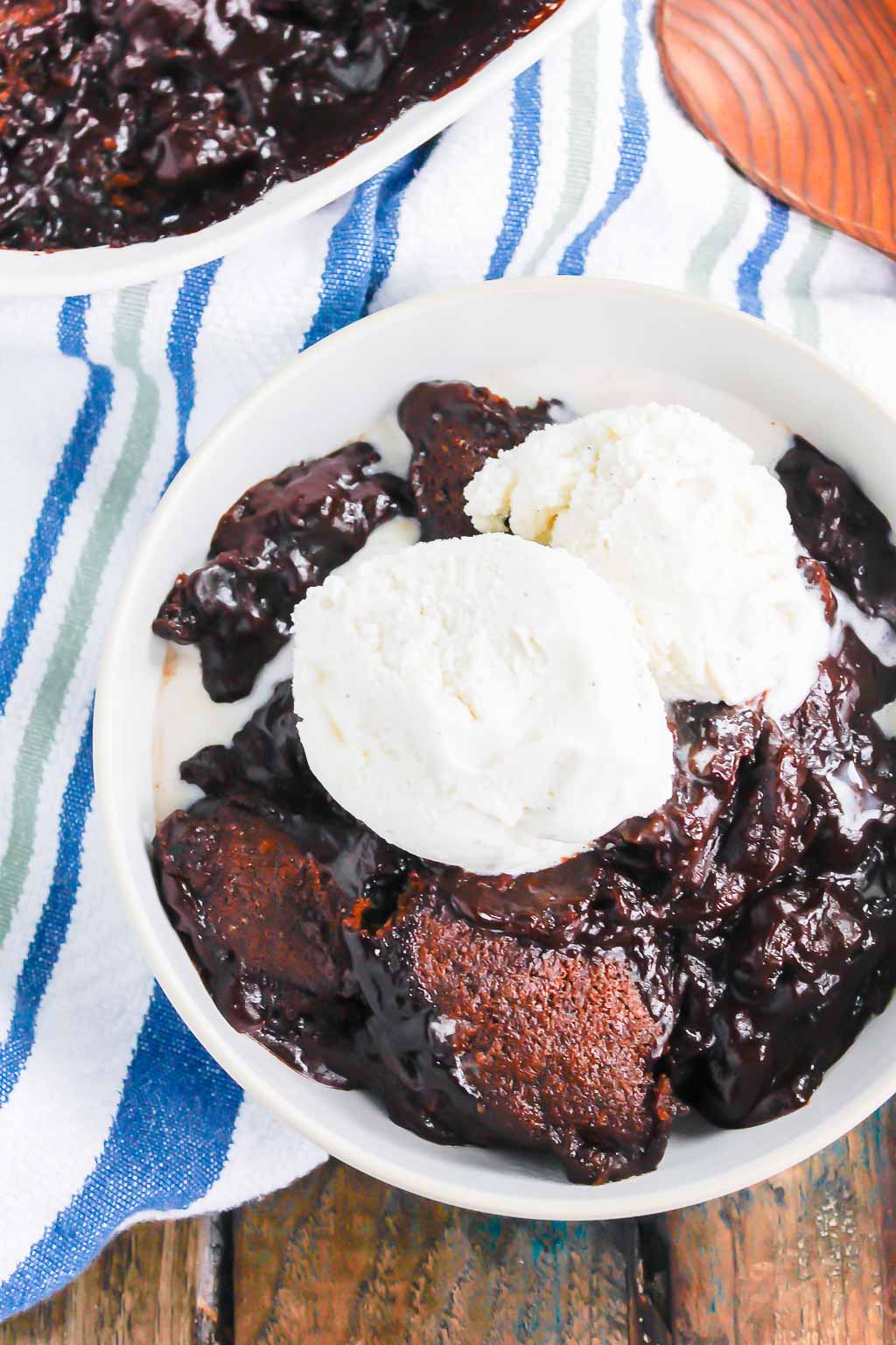 overhead view of serving of chocolate cobbler topped with ice cream in a white bowl. a wooden spoon rests in the background. 