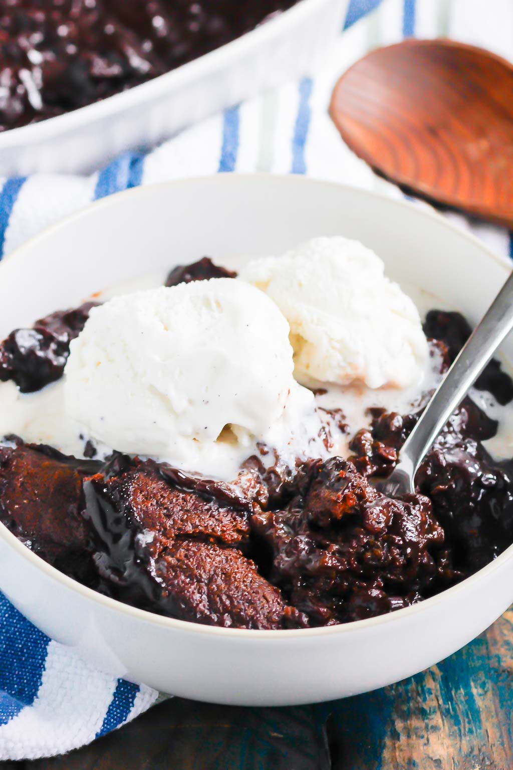 serving of chocolate cobbler topped with ice cream in a white bowl. a wooden spoon rests in the background. 