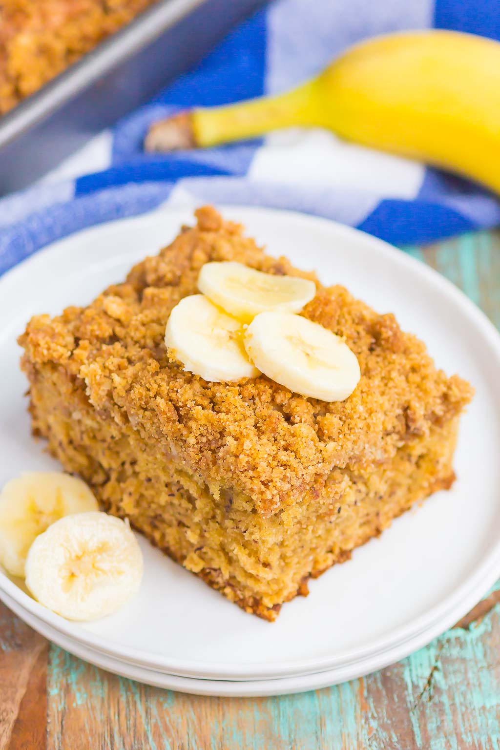 A slice of banana coffee cake topped with banana slices on a white dessert plate. A banana rests on a blue checkered towel in the background. 