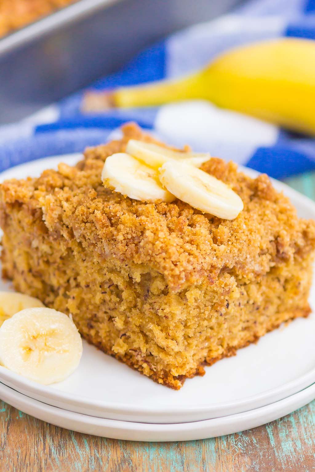A slice of banana coffee cake topped with banana slices on a white dessert plate. A banana rests on a blue checkered towel in the background. 