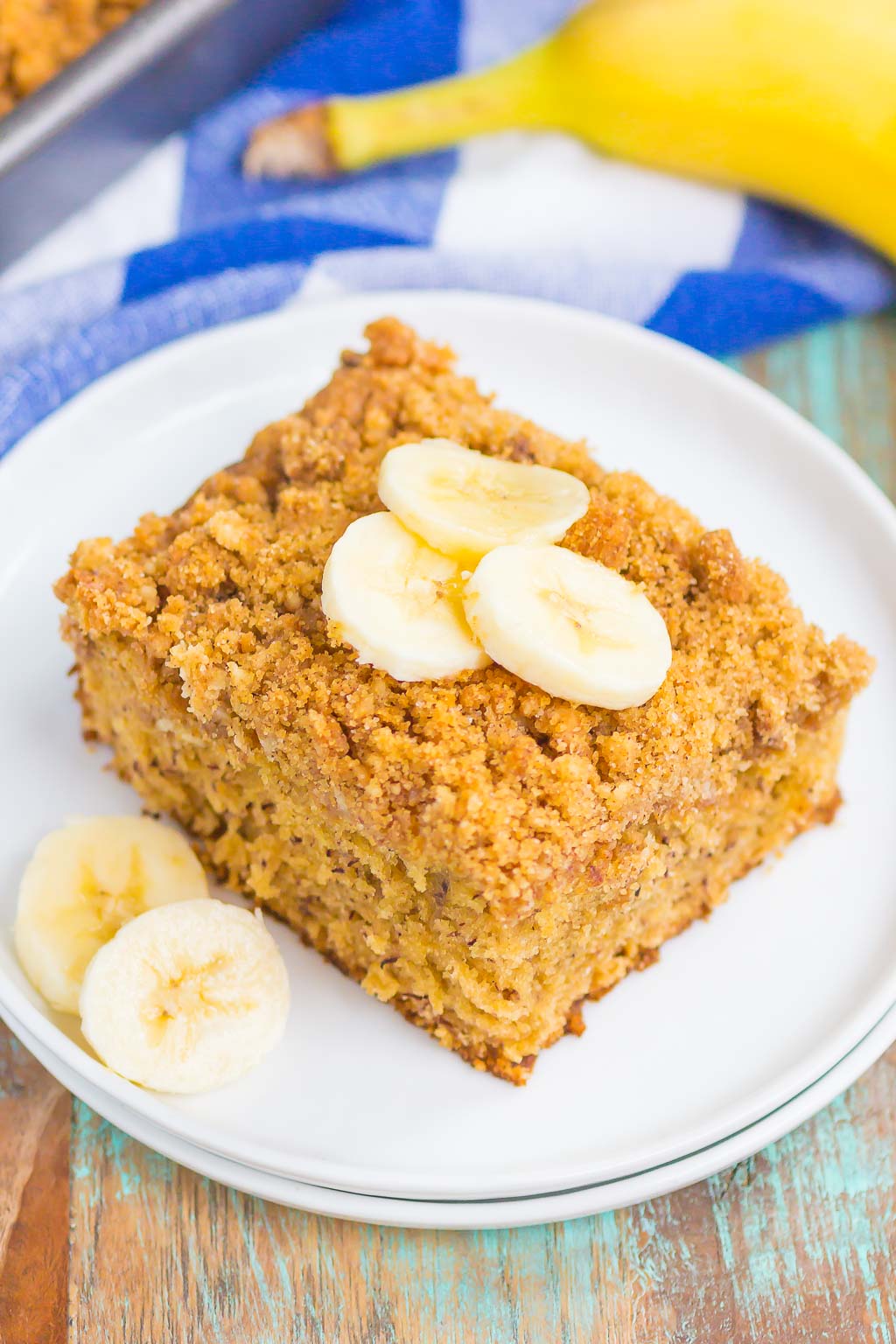 A slice of banana sour cream coffee cake topped with banana slices on a white dessert plate. A banana rests on a blue checkered towel in the background. 