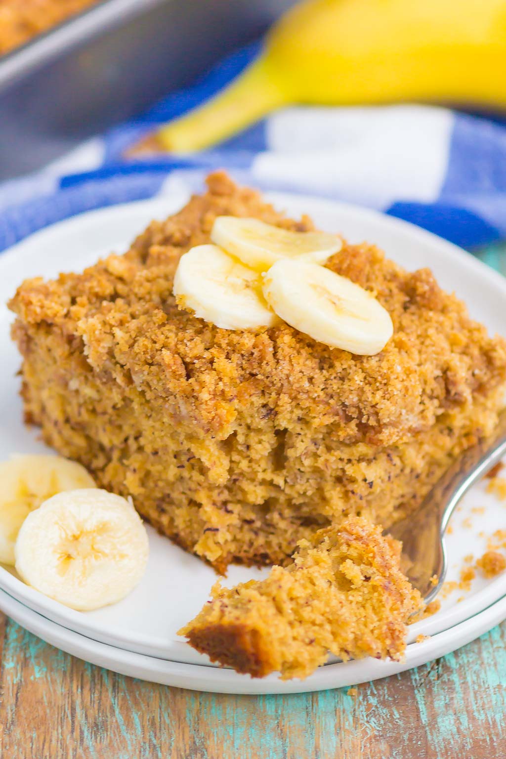 A slice of banana sour cream coffee cake topped with banana slices on a white dessert plate. A banana rests on a blue checkered towel in the background. 