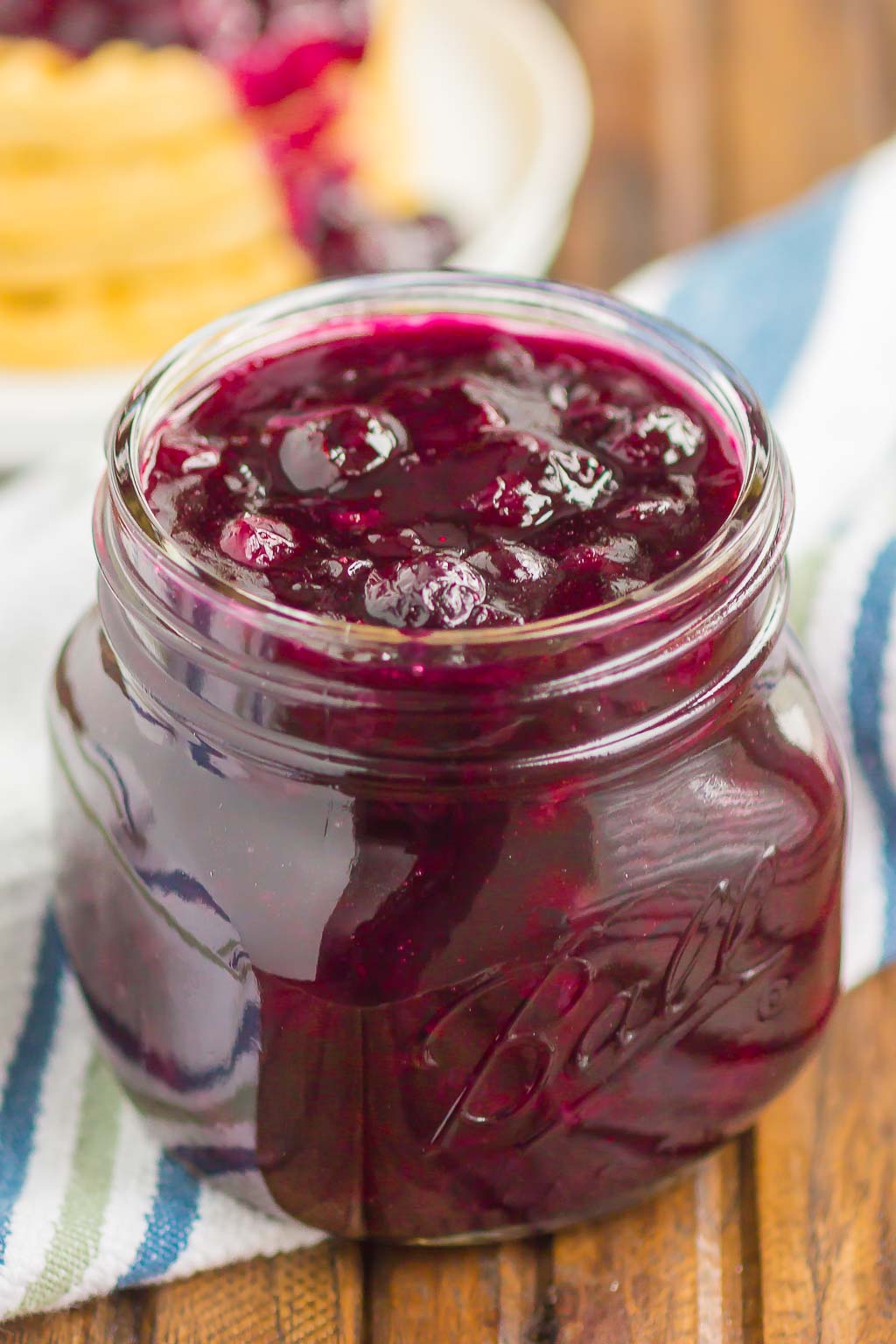 Homemade blueberry sauce in a small ball jar with a stack of waffles in the background. 