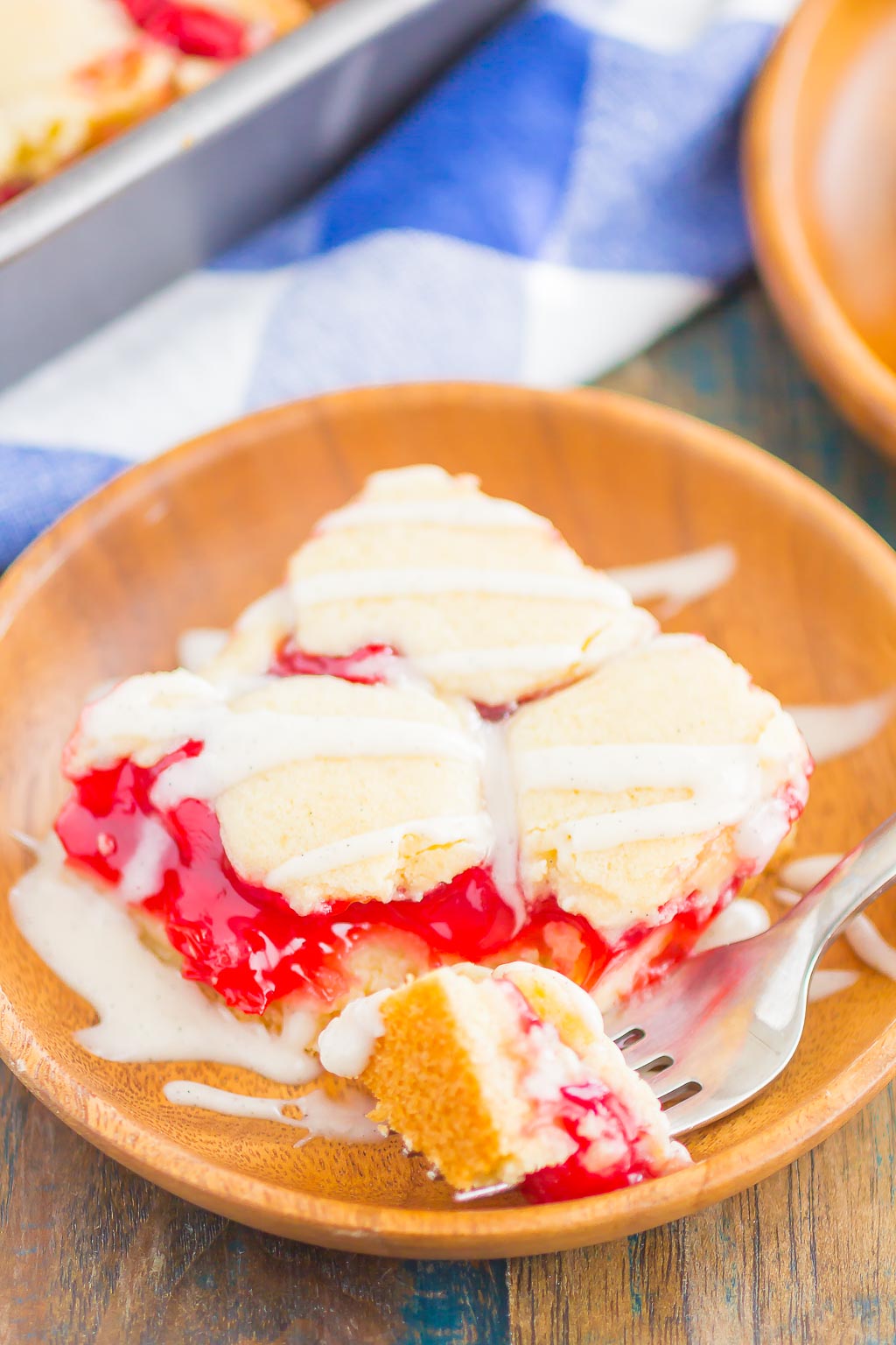 A glazed cherry pie bar on a small wooden plate. A bite is speared on a fork. 