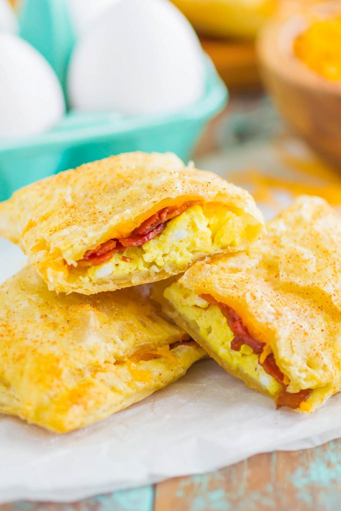A close-up of three breakfast pastries filled with scrambled eggs, bacon, and cheese, stacked on parchment paper. Whole eggs in a teal carton and an orange bowl are blurred in the background.