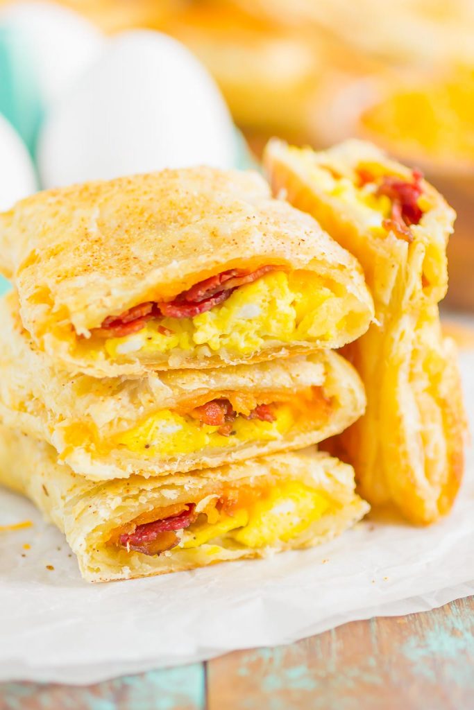 A stack of flaky homemade breakfast hot pockets filled with scrambled eggs, melted cheese, and crispy bacon sits on parchment paper, with two pastries cut open to reveal the savory filling. Whole eggs are blurred in the background.