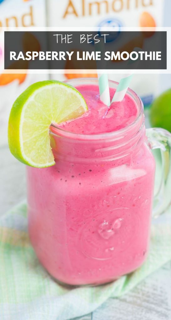 A mason jar filled with a bright pink raspberry lime smoothie, garnished with a lime slice and two blue-striped straws, sits on a cloth with almond milk cartons blurred in the background.
