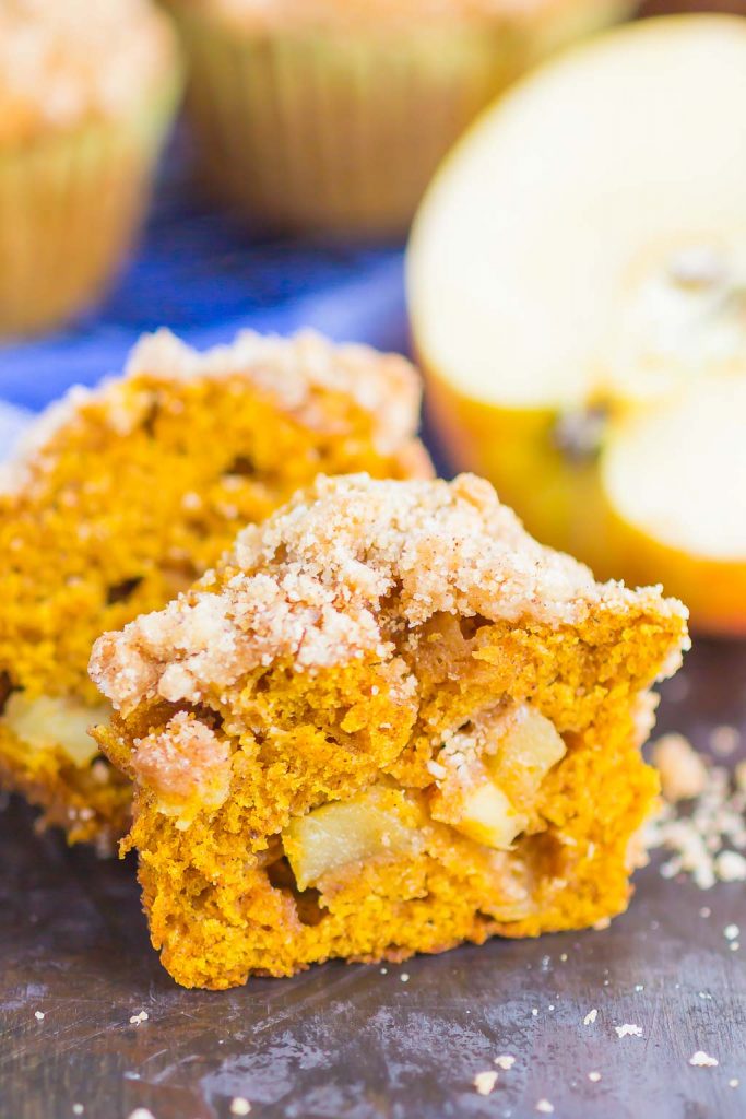 A close-up of a pumpkin apple muffin with a crumb topping, cut in half to reveal apple chunks inside. A sliced apple and blurred muffins are visible in the background.