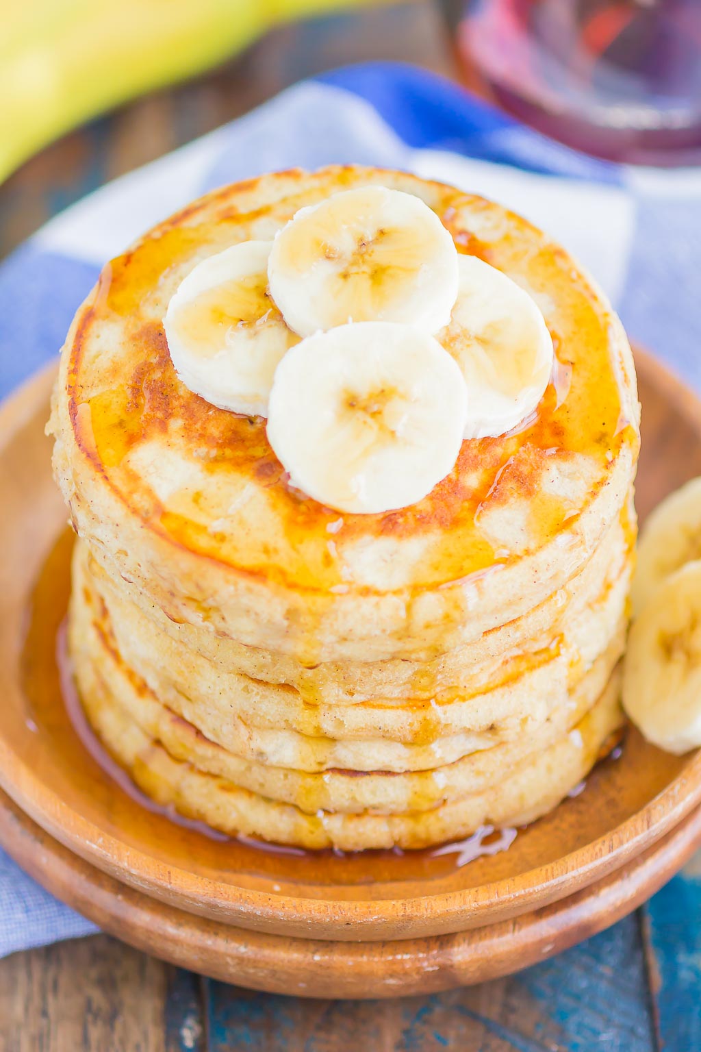 A stack of Banana Pancakes topped with banana slices and maple syrup on a wooden plate.