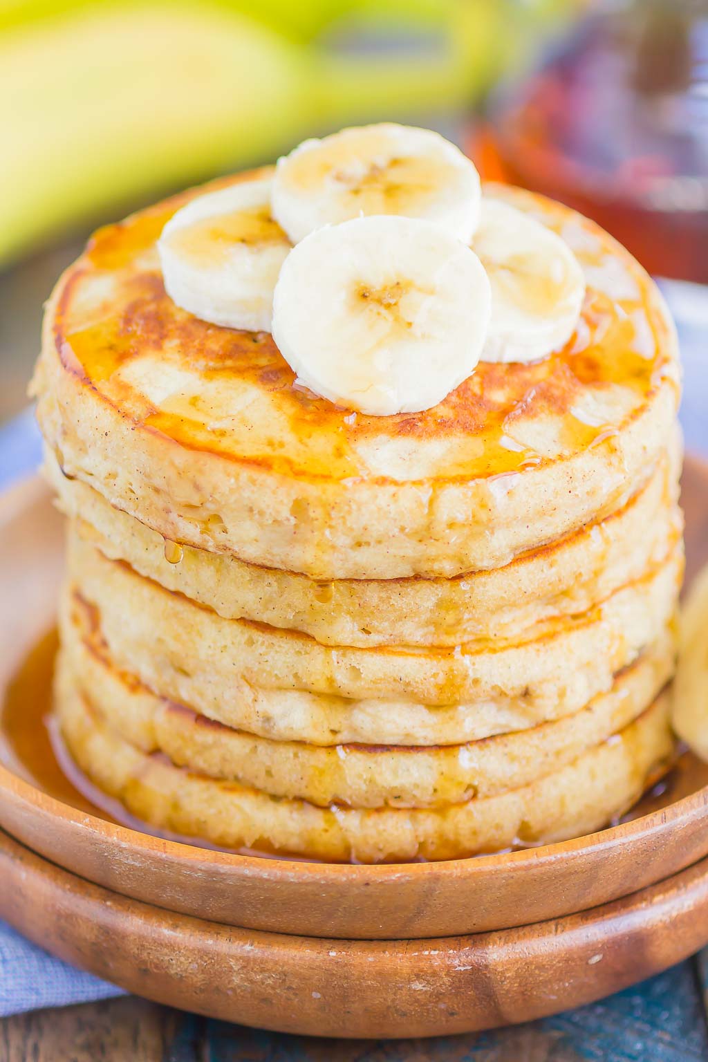 A stack of fluffy Banana Pancakes topped with banana slices and maple syrup on a wooden plate.