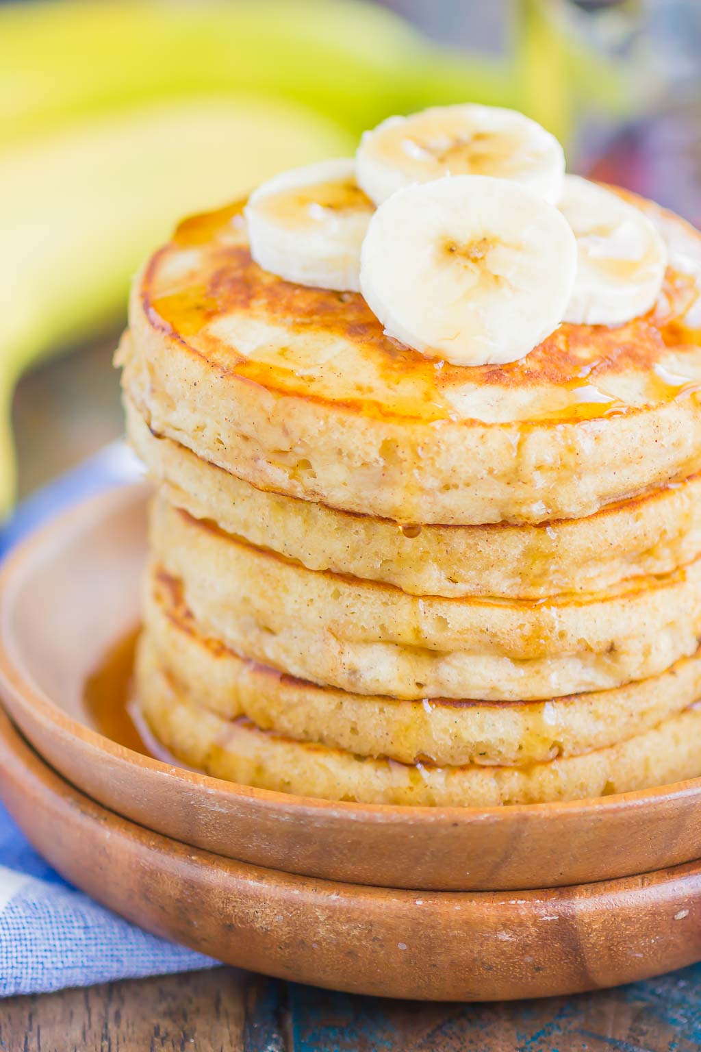 A stack of Banana Pancakes topped with banana slices and maple syrup on a wooden plate.