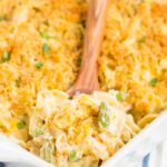 A close-up of a baking dish filled with creamy mac and cheese topped with a golden breadcrumb crust evokes the warmth of chicken and noodle casserole. A wooden spoon scoops out a mix of pasta and vegetables, resting on a striped cloth.