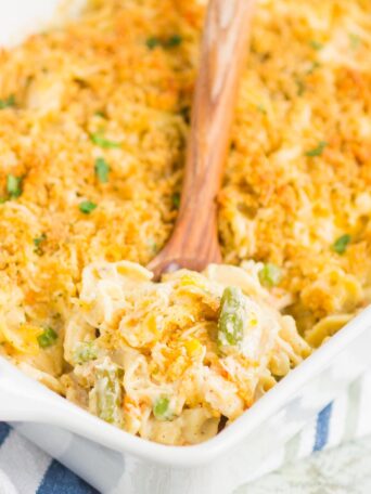 A close-up of a baking dish filled with creamy mac and cheese topped with a golden breadcrumb crust evokes the warmth of chicken and noodle casserole. A wooden spoon scoops out a mix of pasta and vegetables, resting on a striped cloth.