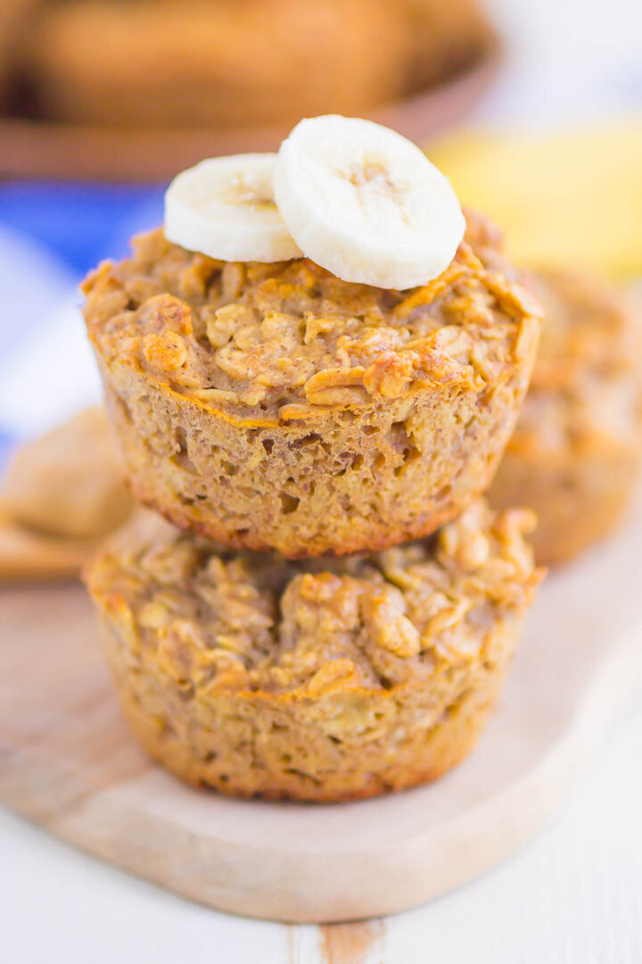 Two peanut butter banana baked oatmeal cups are stacked on a wooden board, with two slices of banana on top. The muffins have a hearty texture, and another baked oatmeal cup is blurred in the background.