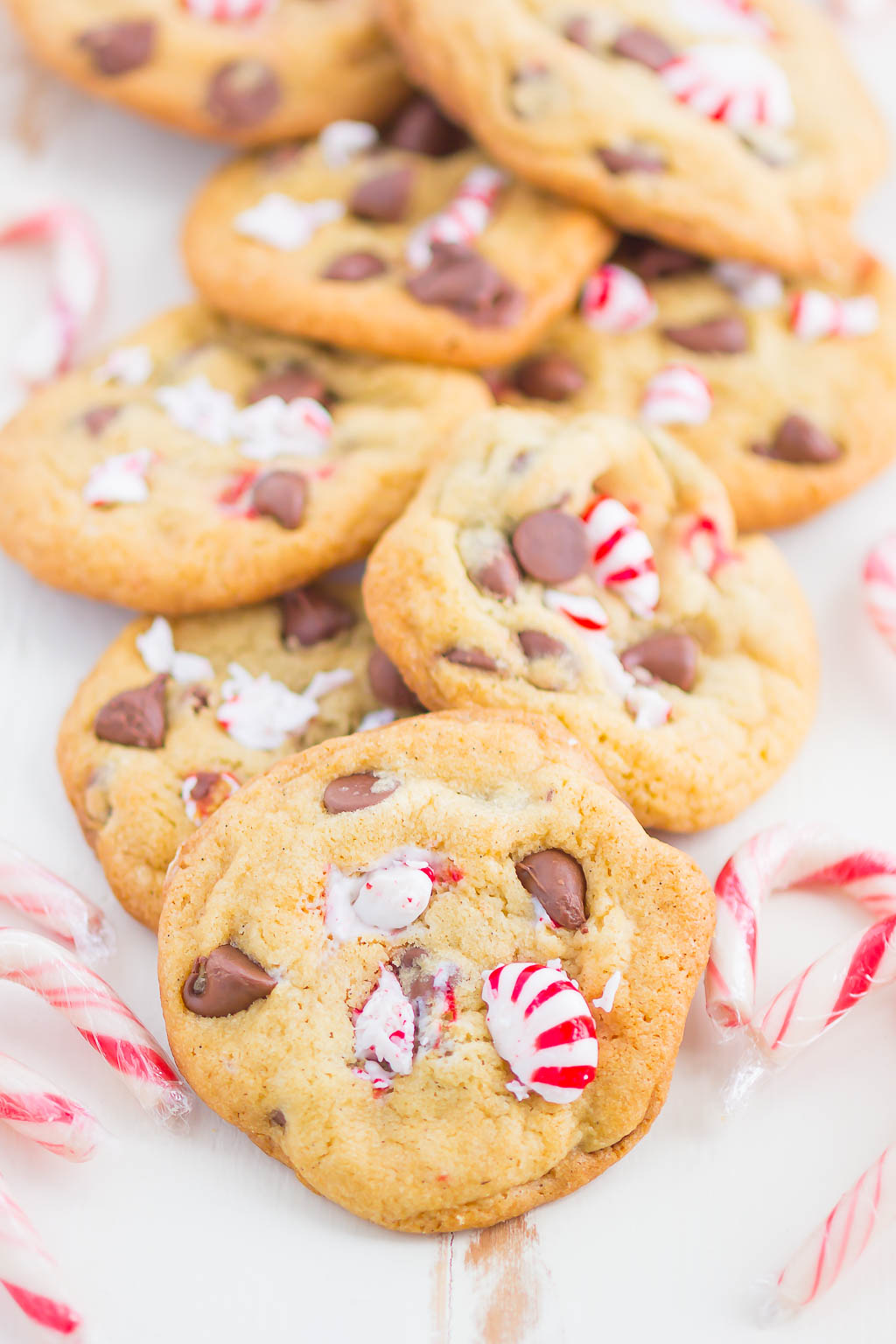 Peppermint Chocolate Chip Cookies piled on a white surface, surrounded by candy canes. 