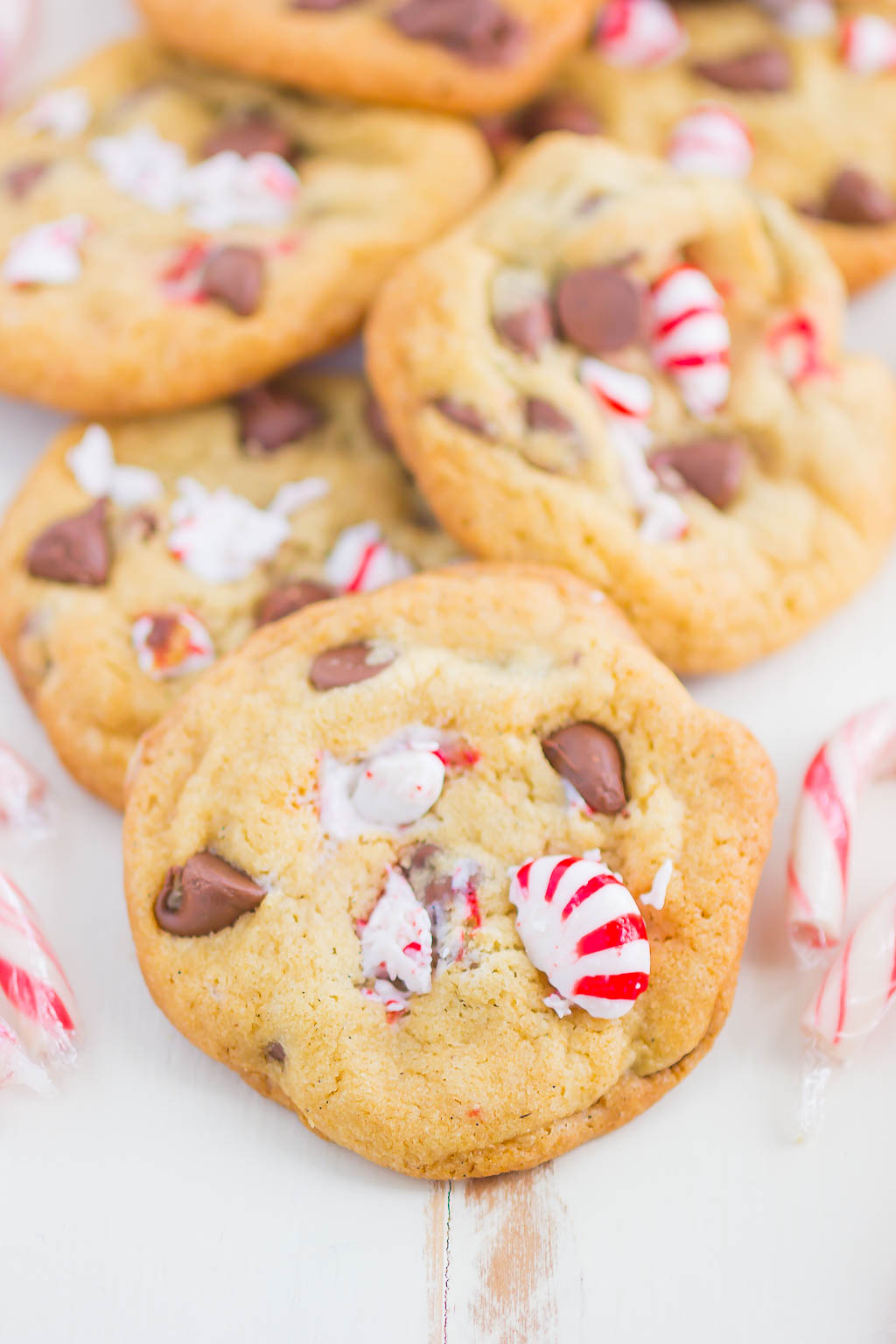 Peppermint Chocolate Chip Cookies piled on a white surface, surrounded by candy canes. 